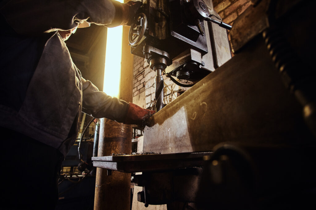 Man is working with giant drill at busy metal factory.